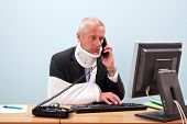 image of health  - Photo of a mature businessman with injuries talking on the phone whilst trying to work on his computer - JPG 