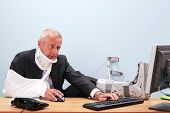 picture of health  - Photo of a mature businessman with multiple injuries sitting at his desk struggling to work on his computer - JPG 
