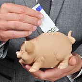stock photo of bill  - closeup of a man wearing a suit introducing a euro bill in a piggy bank - JPG 