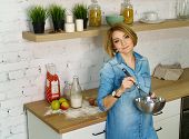 Young attractive girl prepares cookies in the kitchen. She holds a bowl and a confectionery whisk in her hands. Milk, apples, eggs and flour stand on the table